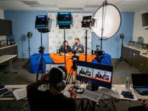 A diverse group of individuals in a room, equipped with cameras and microphones, engaged in a collaborative discussion.